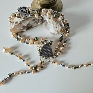 May include: A multi-strand necklace featuring cream, gray, and olive-green pearls. The necklace has a decorative triangular pendant and a silver-tone clasp. The pearls are strung on a dark chain, creating a vintage aesthetic.