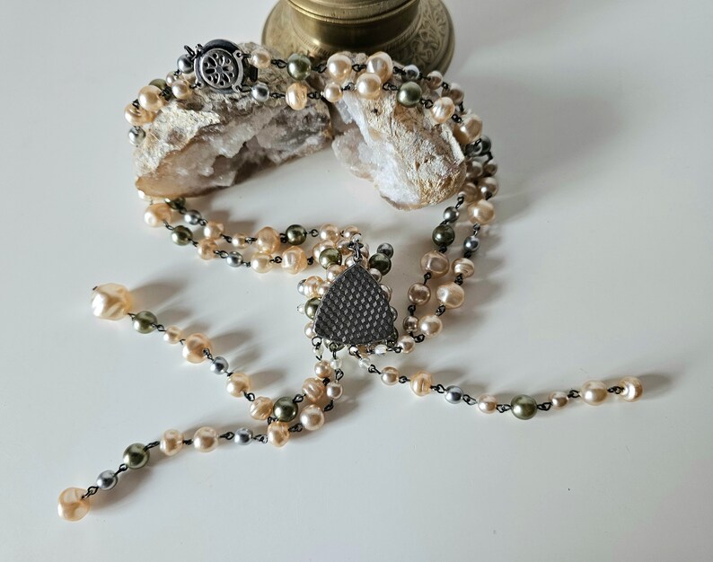 May include: A multi-strand necklace featuring cream, gray, and olive-green pearls. The necklace has a decorative triangular pendant and a silver-tone clasp. The pearls are strung on a dark chain, creating a vintage aesthetic.