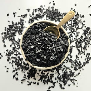 May include: A cream-coloured ceramic bowl filled with black charcoal pieces, with a gold-coloured spoon resting inside. The charcoal is scattered around the bowl on a white surface. The spoon has a decorative design on the handle.