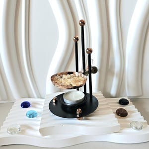 Pode incluir: Um queimador de incenso de metal preto com uma tigela e detalhes em cobre. O queimador contém uma vela de chá acesa e fica em uma plataforma branca ondulada. Pedras decorativas de várias cores circundam a base.