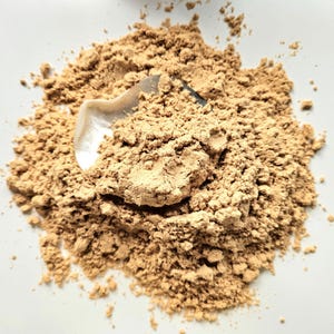 Può includere: Un mucchio di polvere marrone chiaro con un piccolo cucchiaio d'argento parzialmente inserito. La polvere appare finemente macinata e ha una superficie leggermente strutturata, suggerendo un prodotto cosmetico o culinario.
