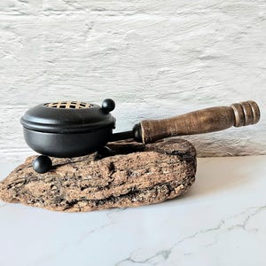 Puede incluir: Un quemador de incienso negro con un mango de madera y una base decorativa de madera. El quemador tiene forma redonda con una parte superior perforada y pequeños pies de bola. El mango es de madera marrón oscuro. La base es un trozo de madera natural.
