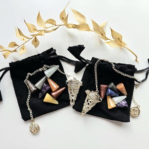 May include: Two black velvet drawstring bags each hold a collection of colorful gemstone pendulums. Each pendulum has a silver chain and a decorative charm. A dried, golden-brown branch with leaves is in the background.