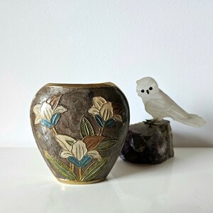May include: A decorative vase with a dark brown background and floral design in white, blue, and brown. A small, white owl figurine sits on a dark rock next to the vase.