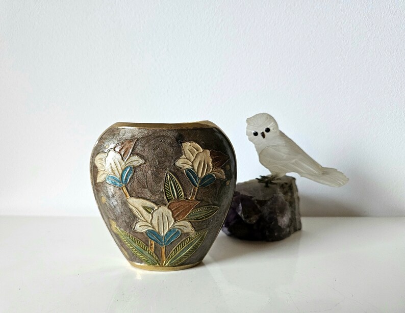 May include: A decorative vase with a dark brown background and floral design in white, blue, and brown. A small, white owl figurine sits on a dark rock next to the vase.