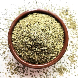 May include: A small, round, brown ceramic bowl filled with dried, chopped green herbs, viewed from above. The herbs are scattered around the bowl on a white surface. The bowl has a simple, unadorned design.