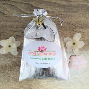 May include: A silver mesh bag tied with a silver ribbon and a gold heart charm. The bag has a white oval label with the text "LOVE VIBRATIONS Incense & Bath Blend Holy Incenses". Two white flowers and a pink stone are next to the bag.