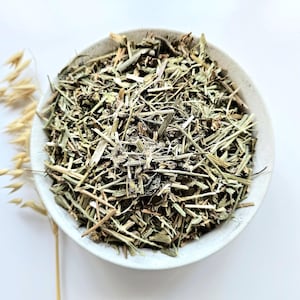 May include: A white ceramic bowl filled with dried green herbs and plant matter. The bowl is set against a white background, with a few stalks of dried wheat to the left. The herbs appear to be cut into small pieces.