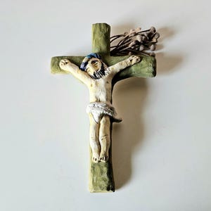 May include: A handcrafted crucifix featuring a figure of Jesus Christ. The cross is painted green with a textured appearance. The figure has a white body, blue hair, and a white loincloth. A bundle of dried plants is attached to the top of the cross.