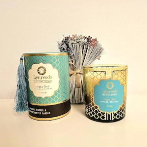 Pode incluir: Duas velas perfumadas com o texto "Ayurveda Aqua Oud" em um design turquesa e dourado. Uma vela é cilíndrica com uma borla, a outra é um frasco de vidro. Um buquê de flores secas está entre elas.