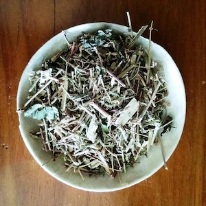May include: A white ceramic bowl containing dried herbs and plant material. The contents are a mix of green, brown, and beige, with visible stems, leaves, and small pieces. The bowl rests on a wooden surface.