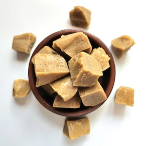 May include: A top-down view of a brown ceramic bowl filled with light brown, square-shaped fudge pieces. Several fudge pieces are scattered around the bowl on a white surface. The fudge has a slightly rough texture.