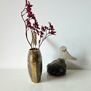 May include: An oval-shaped vase with brass accents and floral designs holds dried red flowers and beige stems. A frosted glass owl figurine sits on a dark purple amethyst crystal, creating a decorative display.