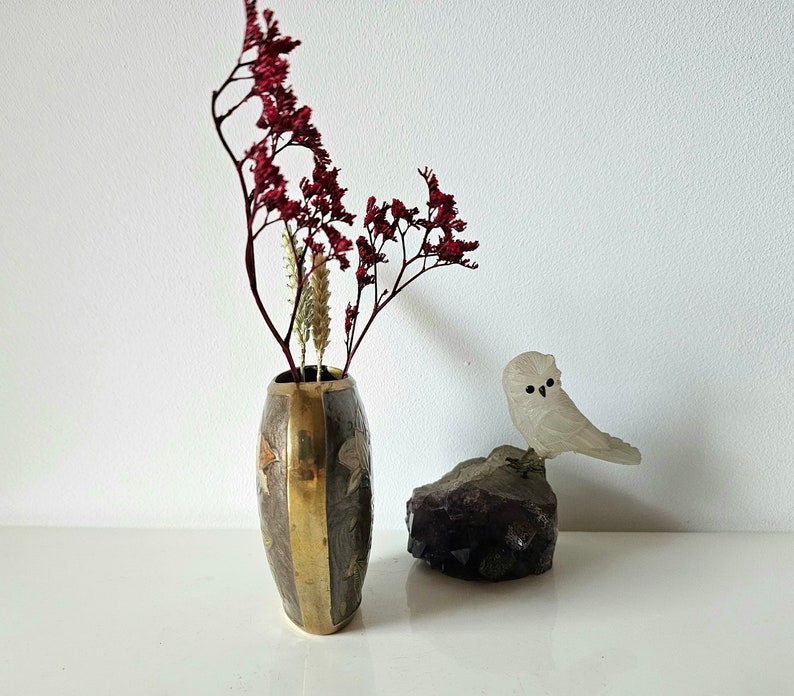 May include: An oval-shaped vase with brass accents and floral designs holds dried red flowers and beige stems. A frosted glass owl figurine sits on a dark purple amethyst crystal, creating a decorative display.