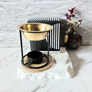 Op de afbeelding: Een goudkleurige oliebrander met een zwarte standaard en een houten basis. Een kleine zwarte beker staat onder de kom. Een witte waxinelichtkaars en een decoratieve vaas met gedroogde bloemen zijn ook aanwezig.