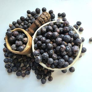 May include: A close-up of dried juniper berries in a white bowl and a small wooden bowl. The berries are dark purple and scattered around the bowls. A wooden scoop is also present.