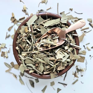 May include: A brown ceramic bowl filled with dried green leaves and stems, with a copper-coloured scoop inside. The scoop and bowl are on a white surface, with some leaves scattered around. The image is a top-down view.