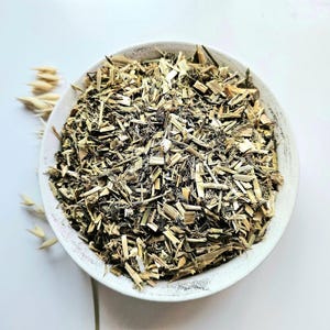 Könnte beinhalten: Eine weiße Keramikschale gefüllt mit getrockneten, gehackten Kräutern. Die Kräuter sind eine Mischung aus hellbraun und grün, mit einer Textur aus kleinen Stücken. Einige Stiele getrockneten Hafers sind links von der Schale auf weißem Hintergrund platziert.