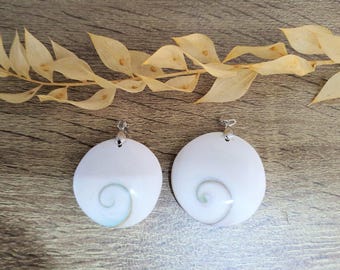 May include: Two white, round pendants with a spiral design in shades of blue and green. Each pendant has a silver-colored bail at the top. The pendants are displayed on a wooden surface with dried leaves.