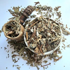 May include: A white ceramic bowl and a small wooden mortar are filled with dried green leaves and stems. The mortar has a carved handle. The leaves are scattered on a white surface. Top-down view.