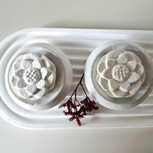 Op de afbeelding: Een witte, decoratieve tray met twee cirkelvormige uitsparingen, elk met een bloemvormig object. Tussen de objecten zit een takje donkerrode gedroogde bloemen. De tray heeft een geribbeld ontwerp op het achterste gedeelte.
