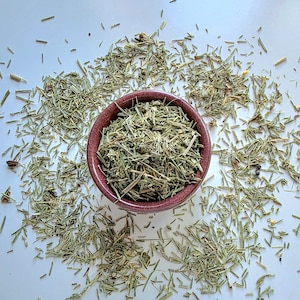 May include: A small, round, dark reddish-brown ceramic bowl filled with dried, light green, needle-like herbal leaves. Additional loose dried herbal leaves are scattered around the base of the bowl, creating a textured, organic arrangement.