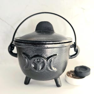 Könnte beinhalten: Ein schwarzer Gusseisentopf mit Deckel und Griff, der auf der Vorderseite ein dreifaches Mondsymbol aufweist. Der Kessel steht auf drei kleinen Beinen. Eine kleine weiße Schale mit Inhalt und eine schwarze Scheibe befinden sich neben dem Kessel.