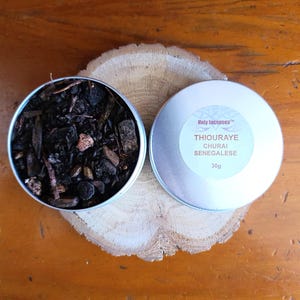 May include: Two small, round, silver tins of incense on a wooden surface. One tin is open, showing dark, textured incense. The other tin is closed, with text that reads "Holy Incenses" and "THIOURAYE CHURAI SENEGALESE 30g".