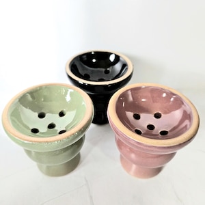 Pode incluir: Três tigelas de cachimbo de água de cerâmica nas cores verde, preto e rosa. Cada tigela tem um design circular com vários orifícios e uma borda bege. As tigelas são empilhadas e têm uma base arredondada.