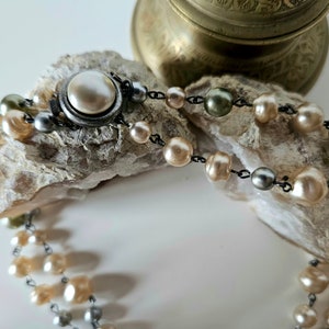 May include: A vintage pearl necklace with a silver clasp. The necklace features multiple strands of cream, gray, and olive-colored pearls. The pearls are strung on a dark chain and rest on a textured, light-colored surface.