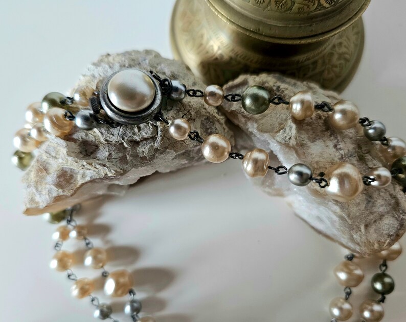 May include: A vintage pearl necklace with a silver clasp. The necklace features multiple strands of cream, gray, and olive-colored pearls. The pearls are strung on a dark chain and rest on a textured, light-colored surface.