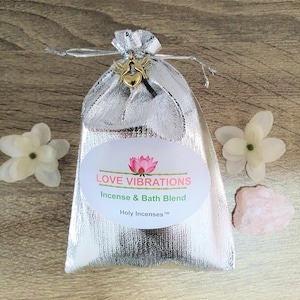 Puede incluir: Una bolsa plateada con cierre de cordón, un dije dorado y una etiqueta que dice "Love Vibrations Incense & Bath Blend". La imagen incluye flores blancas y una piedra rosa, lo que sugiere un tema de spa o bienestar.