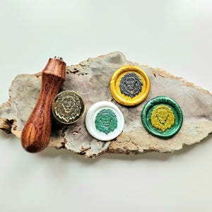 Puede incluir: Un sello de cera de madera con un diseño de león, junto con tres sellos de cera dorados, verdes y blancos, cada uno con un emblema de león. El mango del sello es de color marrón oscuro. Los sellos están sobre un trozo de madera.