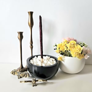 May include: An arrangement featuring a black bowl filled with white stones and a dark red incense stick. Two brass candlesticks and a bat-shaped ring are on display. A white bowl holds yellow and pink flowers.
