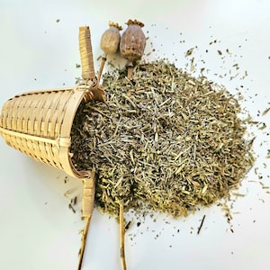 Può includere: Un colino da tè in bambù intrecciato traboccante di erbe essiccate, con due baccelli di semi ed erbe sparse su una superficie bianca. Il colino è marrone chiaro e ha un manico. Le erbe sono un mix di verde e marrone.
