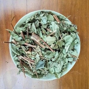 May include: A light green bowl filled with dried green leaves and brown stems, viewed from above. The bowl sits on a wooden surface. The leaves are of various sizes and shapes, with some stems protruding.