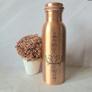 Puede incluir: Una botella de agua de metal color cobre con cuerpo cilíndrico y tapa de rosca. La parte frontal de la botella presenta un gráfico de líneas negras de una flor de loto centrado entre dos diseños ornamentales verticales, simétricos y en espiral.