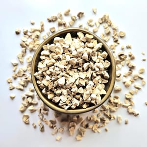 Può includere: Vista dall'alto di una piccola ciotola rotonda marrone piena di pezzi beige chiaro di forma irregolare. La ciotola è circondata da pezzi simili sparsi su una superficie bianca. I pezzi sembrano essere erbe o spezie essiccate.