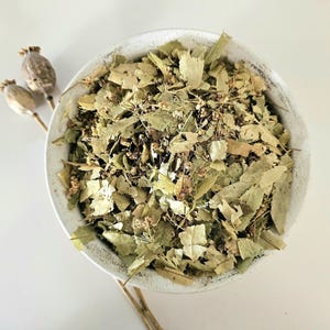 Puede incluir: Un cuenco de cerámica blanco lleno de hojas verdes secas y flores pequeñas. El cuenco está sobre una superficie blanca, con dos vainas de semillas secas y tallos a la izquierda. Las hojas son una mezcla de verde claro y oscuro.