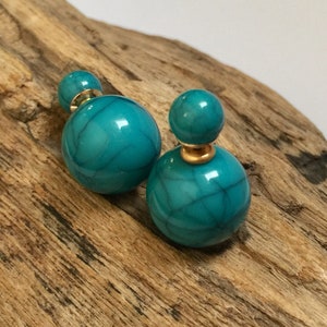Puede incluir: Un par de pendientes de botón turquesa de doble cara. Cada pendiente presenta una cuenta grande y redonda de color turquesa con una cuenta más pequeña de color turquesa en la parte superior, conectadas por un poste dorado. Los pendientes se exhiben sobre madera envejecida.