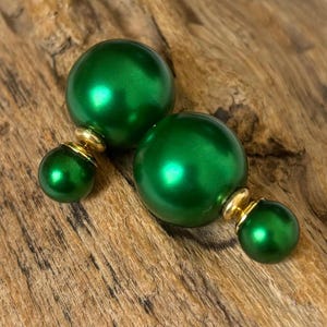 Puede incluir: Un par de pendientes de perlas verdes de doble cara. Los pendientes presentan una perla verde brillante grande y redonda en la parte delantera y una perla verde más pequeña en la parte trasera, conectadas por un poste dorado. Los pendientes se exhiben sobre un trozo de madera.