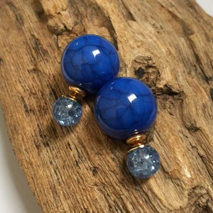 Könnte beinhalten: Ein Paar blaue Kugelohrringe. Jeder Ohrring hat eine große, blau gefärbte Kugel mit Riss-Effekt, die durch ein goldfarbenes Metallstück mit einer kleineren, hellblauen, strukturierten Kugel verbunden ist. Die Ohrringe werden auf einem Stück verwittertem Holz präsentiert.