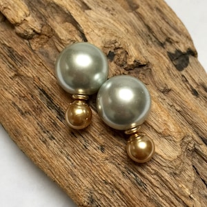 Puede incluir: Un par de pendientes de perlas. Cada pendiente presenta una gran esfera verde grisáceo claro sobre una esfera más pequeña de color dorado. Los pendientes se muestran sobre un trozo de madera marrón claro envejecido.