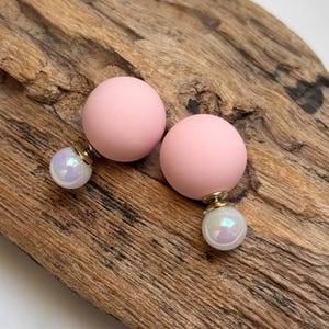 Può includere: Un paio di orecchini a sfera rosa con accenti perlati iridescenti. Gli orecchini sono appoggiati su una superficie di legno marrone chiaro strutturata. Chiusura a perno.