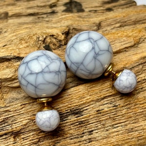 May include: A pair of double-sided stud earrings. Each earring features a large, white marble-patterned sphere with gray veining, connected to a smaller sphere by a gold-colored post. The earrings are displayed on a piece of weathered wood.