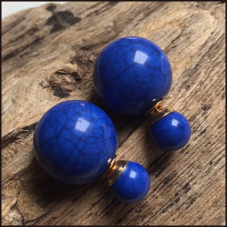 May include: A pair of blue orb earrings with a cracked texture. The earrings have a gold-coloured backing. The earrings are displayed on a piece of wood.