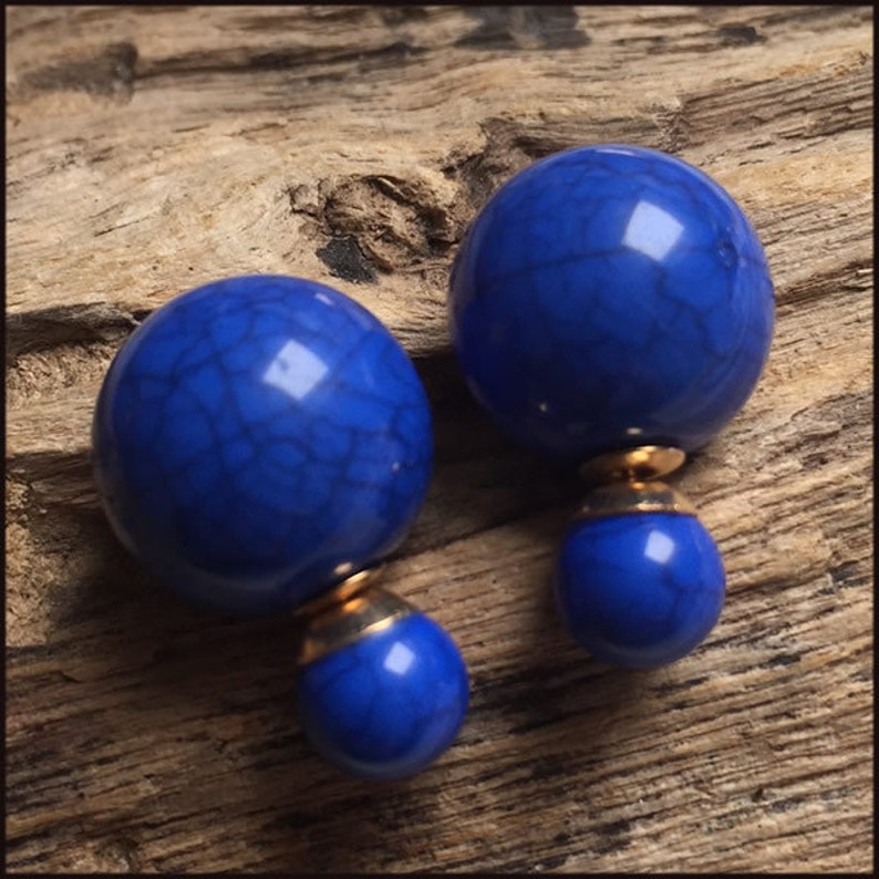 May include: A pair of cobalt blue double-sided earrings. Each earring features a large, round, textured bead with a smaller bead attached below, connected by a gold-coloured metal piece. The earrings are set against a wooden background.