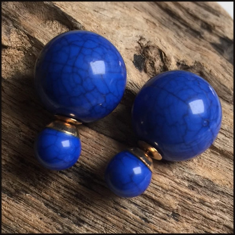 May include: A pair of cobalt blue double-sided earrings. Each earring has a large, round, marbled blue bead attached to a smaller, matching bead with a gold-coloured metal connector. The earrings are displayed on a textured, light brown wooden surface.