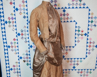 May include: A vintage tan dress on a wooden mannequin. The dress has a high-collared jacket with vertical stripes, a draped bodice, and a pleated skirt. The dress is a historical garment, likely from the late 19th or early 20th century.