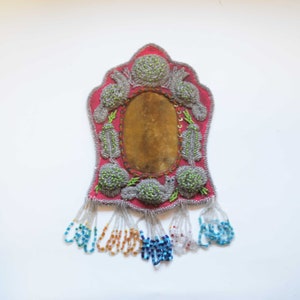 Puede incluir: Un bolso hecho a mano con cuentas, con un fondo rojo y un centro ovalado de color marrón claro. El bolso está adornado con diseños florales de cuentas grises y verdes y flecos de cuentas de varios colores, incluyendo azul, naranja y transparente. El bolso mide aproximadamente 15 cm de altura.